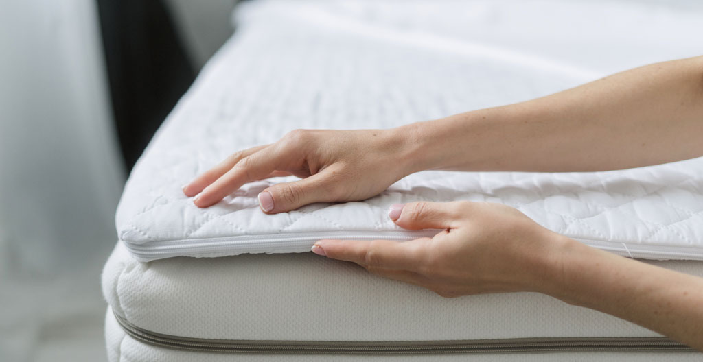 Vue de dos d'une personne ajustant un surmatelas blanc sur un lit dans une chambre moderne lumineuse, geste quotidien naturel